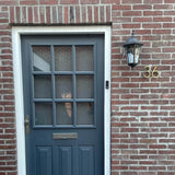 Een donkerblauwe deur met zes glazen ruiten zit in een bakstenen muur. Rechts schitteren een lantaarn en yessential.nl Amato Gold 15cm huisnummers; huisnummer 36 in goud is naast de deur gemonteerd. Eronder zit een brievengleuf.