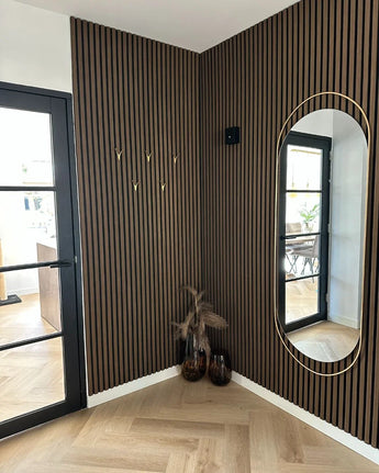 Een moderne entree met verticale houten lattenwanden, een hoge ovale spiegel, minimalistisch design, 10x Vilano Gold gouden kapstokhaakjes van yessential.nl, twee vazen met gedroogde planten, een glazen deur en lichthouten visgraatvloeren.