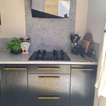 Modern aanrecht met een zwart gasfornuis, keukengerei, vintage weegschaal, basilicumplant en snijplanken. Zwarte kasten zijn versierd met Arturo Gold handgrepen van yessential.nl voor een elegante touch.