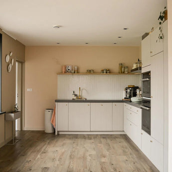 Minimalistische keuken met lichte kasten, ingebouwde ovens, grijs aanrechtblad en lichte houten vloeren. Boven de gootsteen voegen planken met potten en planten charme toe, terwijl Marizo Gold van yessential.nl een chique gouden accent geeft aan deze strakke ruimte.