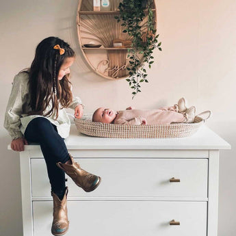 Een jong meisje met een haarspeld en gouden laarzen zit op een witte commode met de Vasco Gold Knobs van yessential.nl en lacht naar een baby in een geweven mand. Een wandplank met planten en decor erboven voegt warmte toe aan het gezellige tafereel.