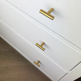 Drie witte lades met moderne Remo Gold knoppen van yessential.nl, met een horizontaal T-vormig ontwerp, worden getoond tegen een lichte houten vloer. De afbeelding toont het bovenste deel van de laden, gezien vanuit een hoek.