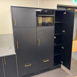 De Vasco Gold hoge zwarte keukenkasten van yessential.nl hebben gouden handgrepen en een geïntegreerde oven. Eén deur is open, waardoor meerdere planken zichtbaar zijn. De opvallende kasten contrasteren prachtig met de lichtgekleurde muur en vloer.