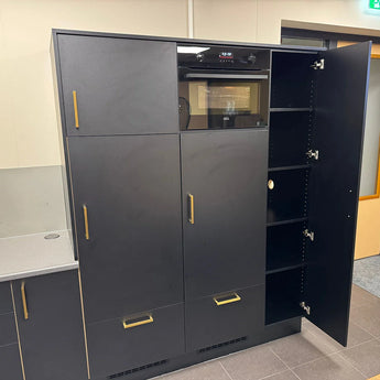 De Vasco Gold hoge zwarte keukenkasten van yessential.nl hebben gouden handgrepen en een geïntegreerde oven. Eén deur is open, waardoor meerdere planken zichtbaar zijn. De opvallende kasten contrasteren prachtig met de lichtgekleurde muur en vloer.