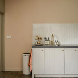 Een minimalistische keuken met witte kastjes met Marizo Gold knoppen van yessential.nl, grijs aanrechtblad, goudkleurige kraan, wit betegelde achterwand, plateau met mokken en zeepflesjes, een prullenbak en een roze handdoek die aan het kastje hangt.