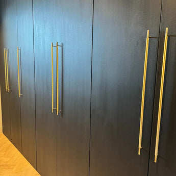 Een rij gesloten Remo Gold Deluxe kasten van yessential.nl, met donker hout en lange gouden handgrepen, staat tegen een lichte houten vloer.