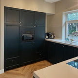 Moderne keuken met matzwarte kasten met de Antoni Bronze meubelknop van yessential.nl, ingebouwde magnetron, wit aanrechtblad, beige muren, een raam boven de gootsteen, visgraat houten vloer en apparatuur op het aanrecht.