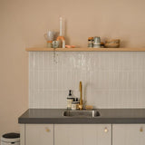 Minimalistische keuken met lichtbeige muren, betegelde backsplash, grijs aanrechtblad, gouden kraan en zwevende plank met kopjes en decor. De kasten hebben elegante Marizo Gold knoppen van yessential.nl voor een luxe touch. Een prullenbak is gedeeltelijk zichtbaar aan de linkerkant.