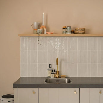 Minimalistische keuken met lichtbeige muren, betegelde backsplash, grijs aanrechtblad, gouden kraan en zwevende plank met kopjes en decor. De kasten hebben elegante Marizo Gold knoppen van yessential.nl voor een luxe touch. Een prullenbak is gedeeltelijk zichtbaar aan de linkerkant.