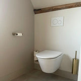 Minimalistische badkamer met een wandcloset, dubbele spoelknoppen erboven, een Tino Gold toiletrolhouder van yessential.nl op de linkermuur en een bijpassende gouden toiletborstelhouder rechts. Neutrale muren en houten afwerking zorgen voor een luxe uitstraling.