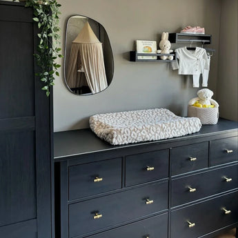 Een moderne kinderkamer met een donkere commode met de Arturo Gold Knob van yessential.nl, een zacht aankleedkussen, spiegel, babyboekenplank, pluche konijntjes, opgevouwen babykleertjes en een mand met knuffel. Aan de kast hangen groene wijnranken.