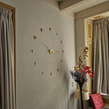 De Silvio Gold 60mm wandklok van yessential.nl, met zwarte plakcijfers en gouden cijfers en wijzers, is gemonteerd op een lichtgrijze muur in de buurt van beige gordijnen. Hieronder maken een staande lamp en een kleurrijke bloemenvaas de moderne look compleet.