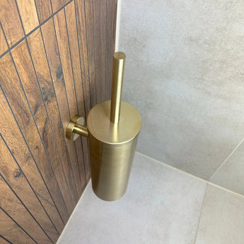 De Branco Gold toiletborstelhouder van yessential.nl is gemonteerd op een wand met bruine houtlook tegels, naast lichtgrijze vloer- en wandtegels in een badkamerhoek.