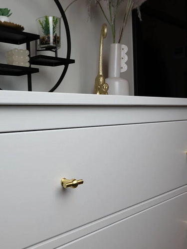Een witte commode met 10x Rando Gold Knob van yessential.nl voegt elegantie toe, met daarop vazen en een moderne zwarte ronde plank waarop kleine planten en ornamenten staan.
