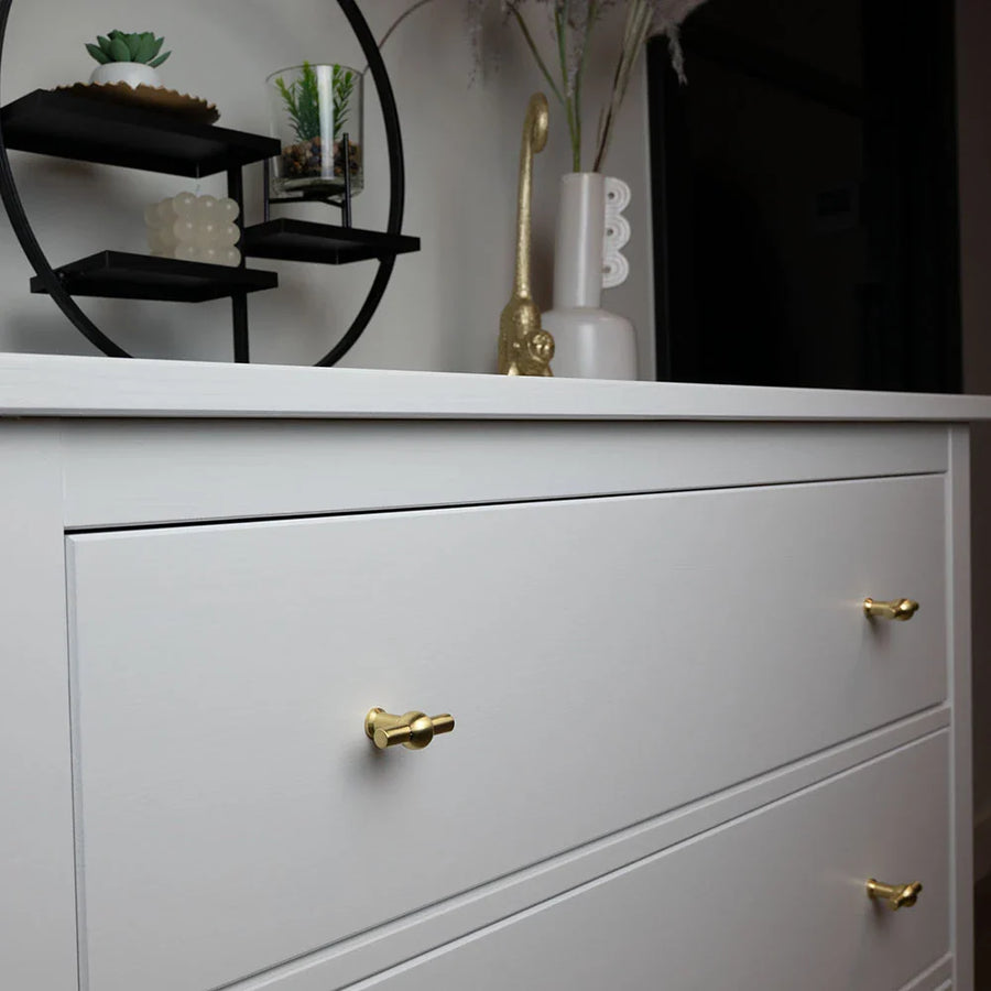 Een witte commode met 10x Rando Gold Knob van yessential.nl voegt elegantie toe, met daarop vazen en een moderne zwarte ronde plank waarop kleine planten en ornamenten staan.