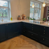 Moderne keuken met donkerblauwe kasten met Antoni bronzen knoppen van yessential.nl, witte werkbladen, houten keukengerei, planten in potten en grote ramen met uitzicht op bomen. Warme visgraat houten vloeren maken de ruimte compleet.