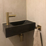 Een kleine zwarte rechthoekige wastafel met een moderne gouden kraan is gemonteerd in de badkamerhoek, aangevuld met yessential.nl's Damiano Gold WC-set. Een beige handdoek hangt aan een goudkleurig haakje aan de betegelde muur.