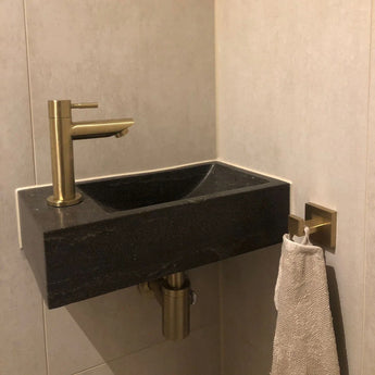 Een kleine zwarte rechthoekige wastafel met een moderne gouden kraan is gemonteerd in de badkamerhoek, aangevuld met yessential.nl's Damiano Gold WC-set. Een beige handdoek hangt aan een goudkleurig haakje aan de betegelde muur.