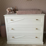 Een witte commode met drie lades, luxe gouden afwerking en yessential.nl Vasco Gold Knob handgrepen is bedekt met een roze aankleedkussen, knuffels en babyverzorgingsspullen, tegen een muur met textuur en patronen.