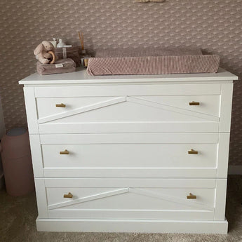 Een witte commode met drie lades, luxe gouden afwerking en yessential.nl Vasco Gold Knob handgrepen is bedekt met een roze aankleedkussen, knuffels en babyverzorgingsspullen, tegen een muur met textuur en patronen.