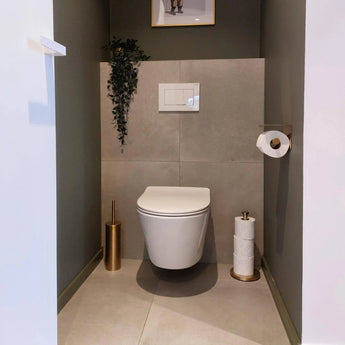 Een moderne, minimalistische badkamer met de Tygo Gold WC Set (no-drill) van yessential.nl: bevat een wandcloset, gouden toiletborstel met houder, reserverolhouder en een slepende plant boven de spoelplaat op een betegelde muur.