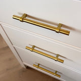 Drie witte lades met moderne Arturo Gold handgrepen van yessential.nl, getoond vanuit een bovenliggende hoek. De lichtbruine houten vloer eronder complementeert het minimale, eigentijdse ontwerp.