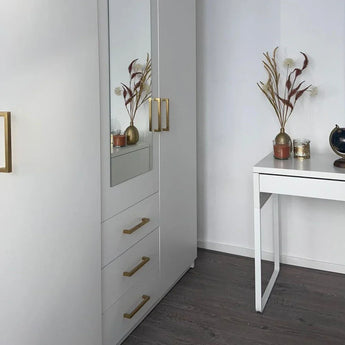 Een moderne, minimalistische kamer met witte meubels, met de yessential.nl Vasco Gold kledingkast met gouden handgrepen en een spiegel, plus een klein bureau versierd met vazen met gedroogde bloemen en een wereldbol op een donkerhouten vloer.