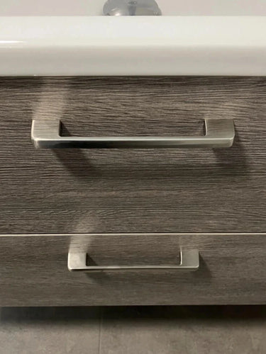 Een close-up van twee Ramiro Chrome Deluxe handgrepen van yessential.nl op een lade van een badkamermeubel met houtstructuur, met een witte wasbakrand erboven en strakke grijze tegels op de vloer.