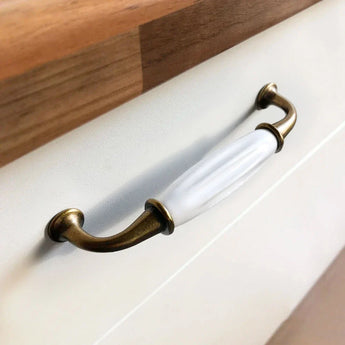 Een close-up van de Daro Ceramic Antique Gold handgreep van yessential.nl, met antiek goud en wit keramiek op een lade met een houten bovenkant - ideaal voor fans van klassiek meubelbeslag.