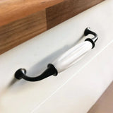 Close-up van de Daro Ceramic Black handgreep van yessential.nl, met een zwart metalen uiteinde en witte keramische greep, gemonteerd op een wit ladefront onder een houten aanrechtblad.
