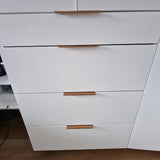 Een close-up van vier witte kastladen met yessential.nl Enzo Rose Gold moderne meubelgrepen in een horizontale afwerking, naast een gedeeltelijk geopende bijpassende kastdeur, alles op een houten vloer.