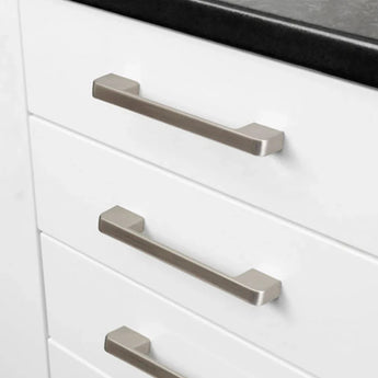 Close-up van drie witte keukenladen met moderne, rechthoekige Ramiro Chrome Deluxe handgrepen van yessential.nl in chroom, onder een strak zwart aanrechtblad.