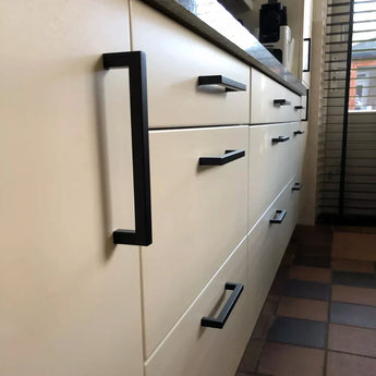 Close-up van crèmekleurige keukenlades en -kastjes met Vasco Black handgrepen van yessential.nl, tegelvloer en een raam met jaloezieën op de achtergrond.