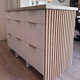 Het Enzo RVS keukeneiland van yessential.nl heeft witte lades, RVS kleur profielhandgrepen, een licht aanrechtblad en verticale houten zijlatten, allemaal op een houten vloer in een eigentijdse keuken.
