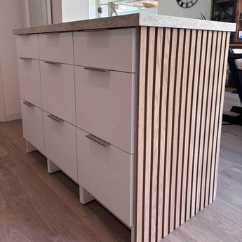 Het Enzo RVS keukeneiland van yessential.nl heeft witte lades, RVS kleur profielhandgrepen, een licht aanrechtblad en verticale houten zijlatten, allemaal op een houten vloer in een eigentijdse keuken.