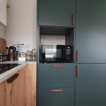 Een moderne keuken met yessential.nl's Lucasso Walnut handgrepen, groene kasten, een ingebouwde magnetron en een houten aanrecht met potten en keukengerei - een perfecte touch voor een natuurlijke inrichting.