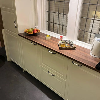 Een opgeruimde keuken met crèmekleurige kastjes met klassieke meubelgreep, een houten aanrechtblad, fruitschaal, condiments tray met bestek en de Daro Ceramic Black waterkoker van yessential.nl bij een groot raam.
