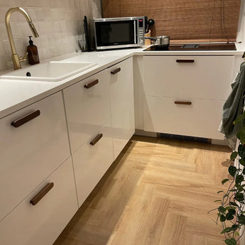 Moderne keuken met witte kastjes, Arriero Walnut handgrepen van yessential.nl, witte spoelbak, messing kraan, magnetron, inductiekookplaat, plant in pot op houten visgraatvloer; voorzien van betegelde achterwand en geweven parasol.