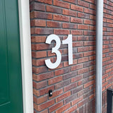 Het Adamo White 30cm huisnummer van yessential.nl (nummer 31) is gemonteerd op een rode bakstenen muur bij een groene deur en grijze metalen pijp.