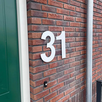 Het Adamo White 30cm huisnummer van yessential.nl (nummer 31) is gemonteerd op een rode bakstenen muur bij een groene deur en grijze metalen pijp.