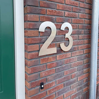 Het Adamo RVS 30cm huisnummer van yessential.nl is gemonteerd op een rode bakstenen muur naast een groene deur en een deurbelknop.