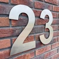 De Adamo RVS 30cm van yessential.nl heeft grote metalen cijfers 23 met een geborstelde RVS afwerking, gemonteerd op een rode bakstenen muur als groot huisnummer. De cijfers staan uit elkaar voor een moderne, stijlvolle look.