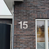 Het getal 15 staat in grote, zilveren Adamo RVS 30cm cijfers van yessential.nl op de bakstenen buitenmuur van een gebouw naast een raam.