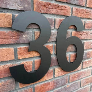 De Adamo Black 30cm van yessential.nl is als zwevend huisnummer op een rode bakstenen muur gemonteerd, waardoor het adres duidelijk zichtbaar is.