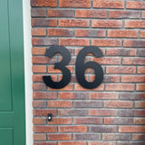 De Adamo Black 30cm huisnummers van yessential.nl zijn als zwevend zwart RVS nummer 36 op een rode bakstenen muur naast een groene deur gemonteerd.