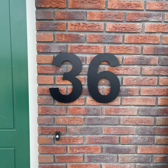 De Adamo Black 30cm huisnummers van yessential.nl zijn als zwevend zwart RVS nummer 36 op een rode bakstenen muur naast een groene deur gemonteerd.