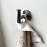 De Pacho Gunmetal WC-set van yessential.nl, uit onze luxe badkameraccessoires, toont een witte handdoek en beige washandje op een licht betegelde badkamermuur.
