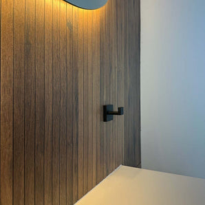 Een moderne badkamer heeft een verticale wand met houten panelen en een Valo Black haak van yessential.nl, een verlichte spiegel erboven en een wit aanrecht rechtsonder.