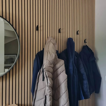 Vier jassen hangen aan de yessential.nl 10x Huso Black haken die bevestigd zijn aan een verticale houten lattenwand. Links weerspiegelt een ronde spiegel met een zwarte lijst gedeeltelijk de nette, moderne ruimte.