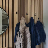 Een houten lattenwand is voorzien van vier yessential.nl Huso Black haken, die elk een beige of blauwe jas of jack vasthouden. Aan de linkerkant van de muur hangt een ronde spiegel.